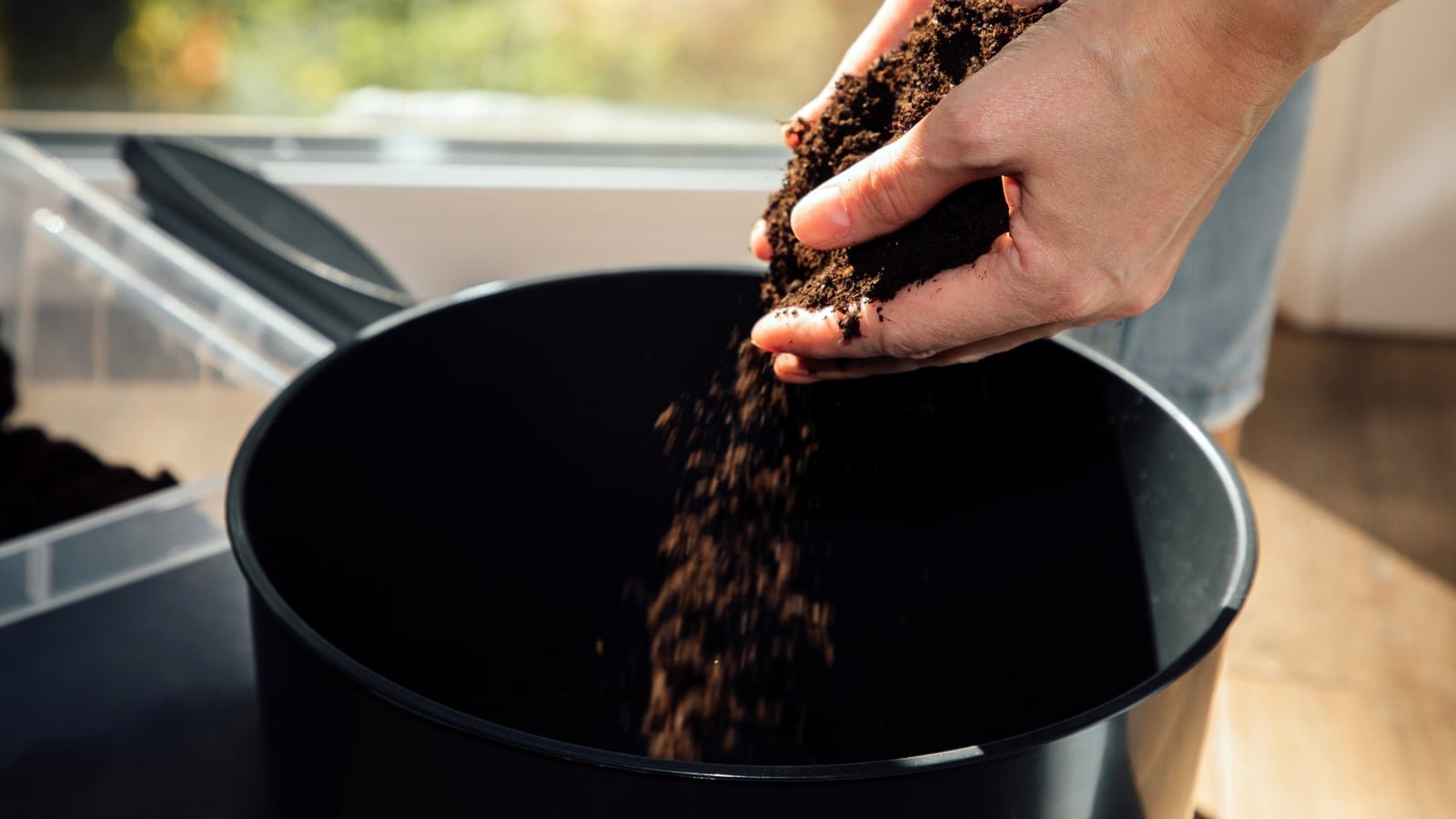 Female hands pouring fresh loose black soil into a large black plastic pot on a sunny balcony.
